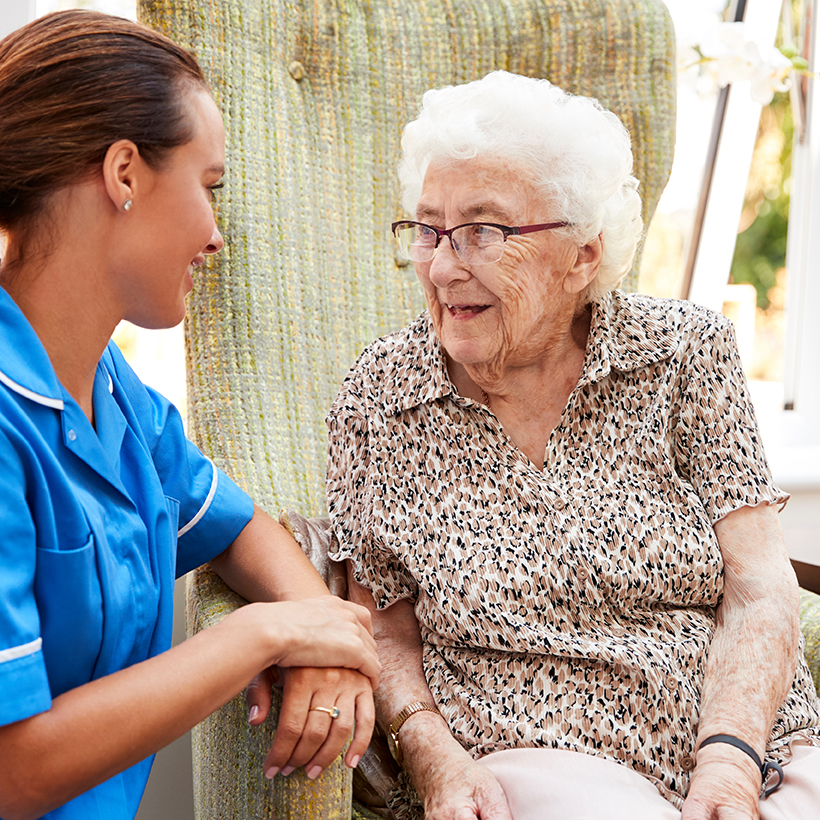 Senior Woman Sitting In Chair And Talking With Nurse In Retirement Home